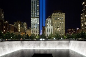 The Tribute in Light rises behind buildings adjacent to the World Trade Center complex and a reflecting pool at the National September 11 Memorial, Monday, Sept. 8, 2014, in New York. The tribute, an art installation of 88 searchlights aiming skyward in two columns, is a remembrance of the Sept. 11, 2001, attacks. (AP Photo/Mark Lennihan)