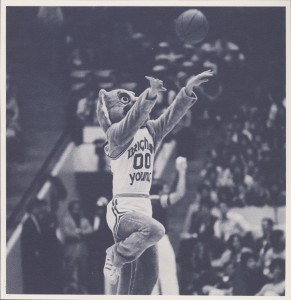 Cosmo takes a half-court shot during a game in the 1980-81 basketball season. Cosmo is an entertainer at any appearance he makes. (L. Tom Perry Special Collection)